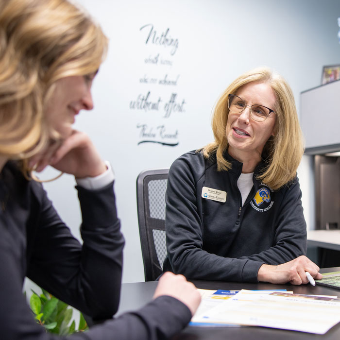 A staff member in an office hands a college folder across the desk to another person during an advising-style meeting.
