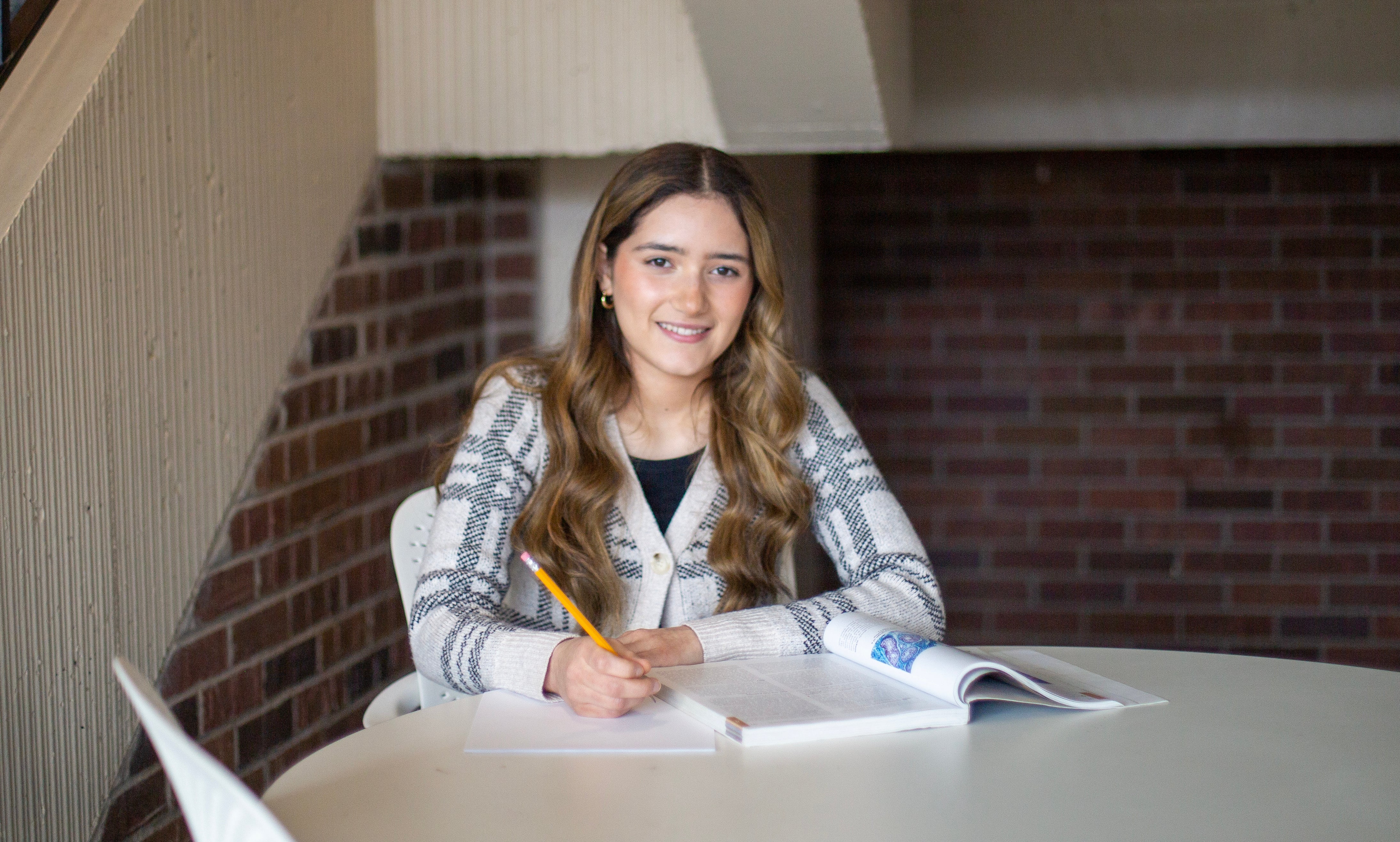 Female student sits at table on campus holding a pencil writing in notebook