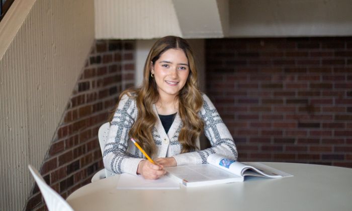 Female student sits at table on campus holding a pencil writing in notebook