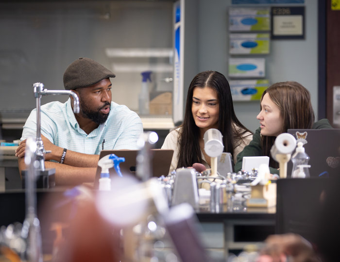 One male student and two female students in science lab talking with various lab equipment in foreground