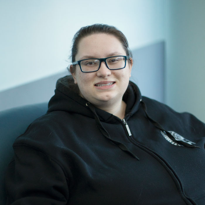 Student seated indoors, wearing glasses and a dark hoodie, looking toward the camera in a campus lounge or hallway setting.
