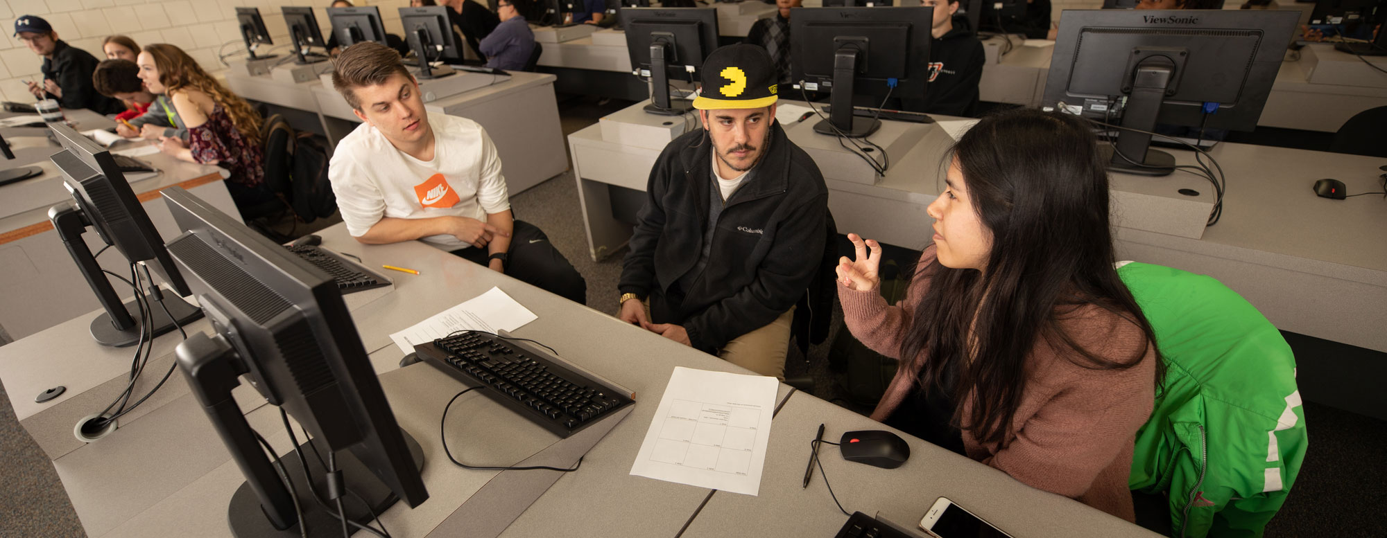 Group of students seated at computer workstations discussing a class assignment in a computer lab
