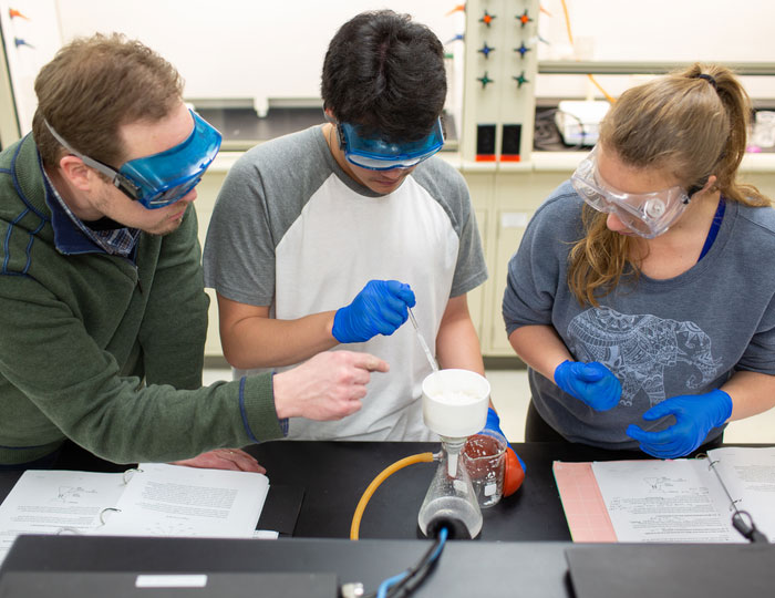 Students and an instructor collaborate on a chemistry lab experiment using a vacuum filtration setup, wearing gloves and safety goggles at a laboratory bench.
