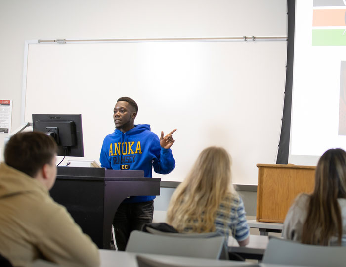 male in blue sweatshirt in front of class pointing at screen with presentation