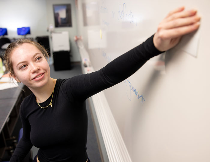 Person writing on a whiteboard in a classroom with desks and computers nearby.