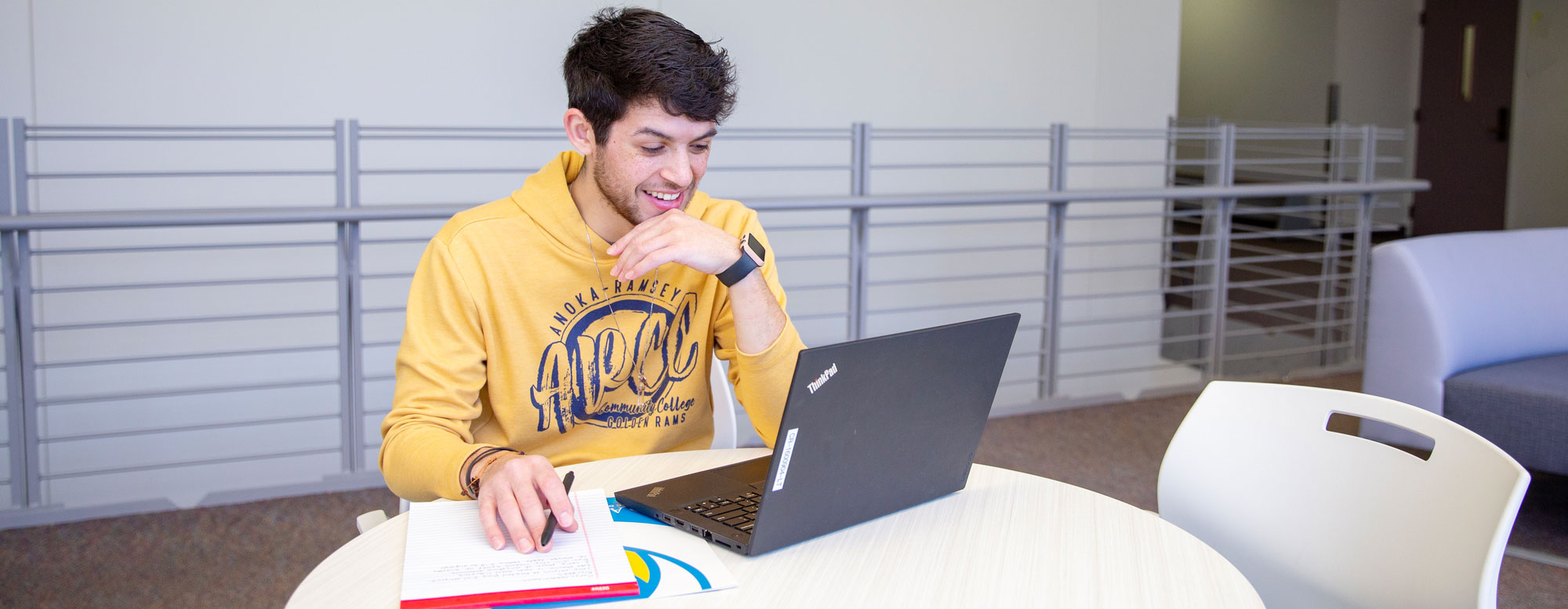 male in yellow anoka-ramsey sweatshirt smiling at computer with writing pad