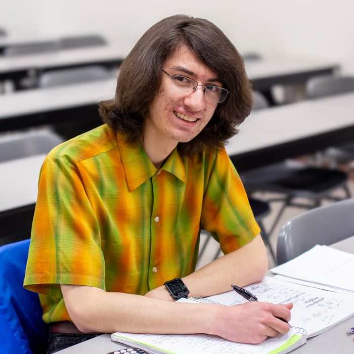 Student taking notes in a notebook at a classroom desk with lecture seating in background