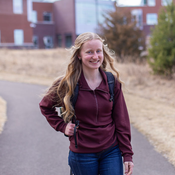 smiling student walking on path outside campus with one hand on backpack