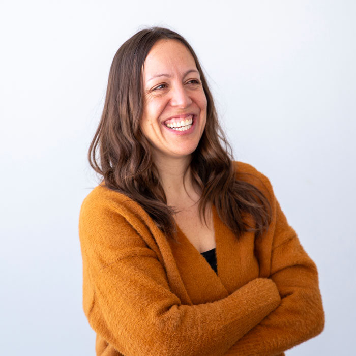 Portrait-style photo of a person with long brown hair, arms crossed, wearing a brown sweater, standing against a light background.
