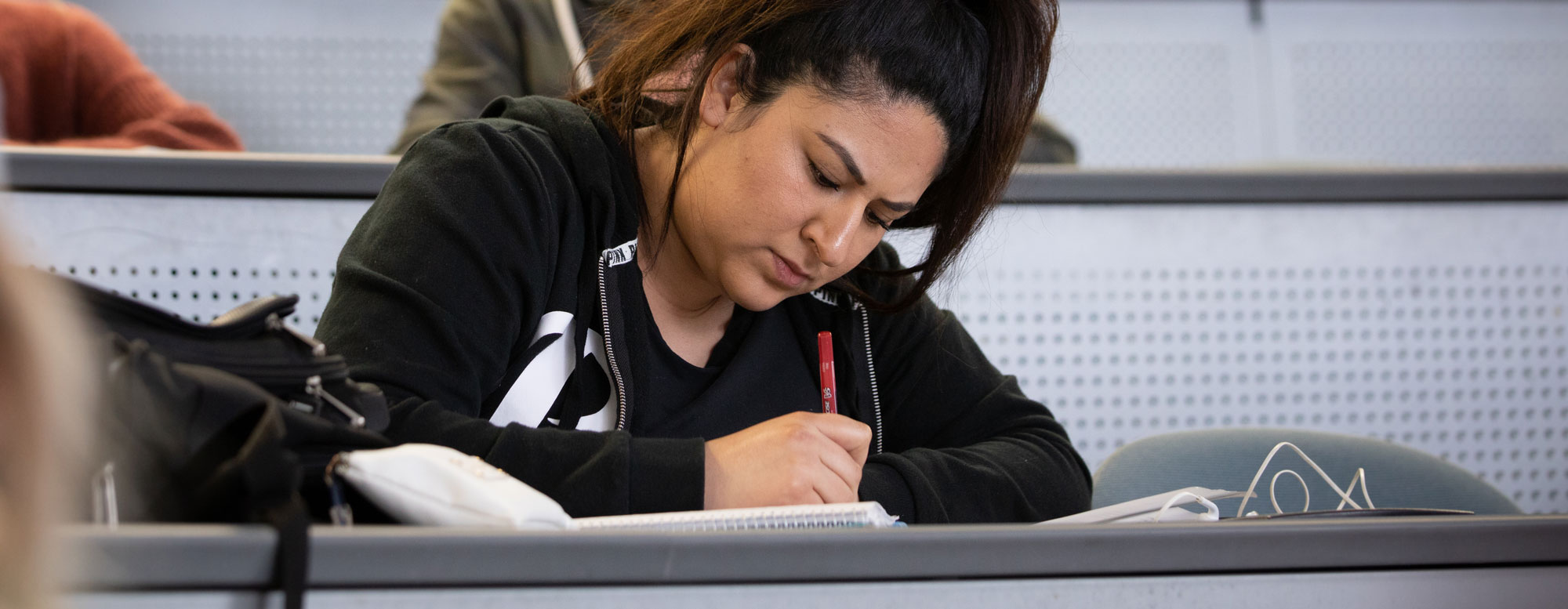 College student writing notes during a lecture in a classroom with tiered seating.