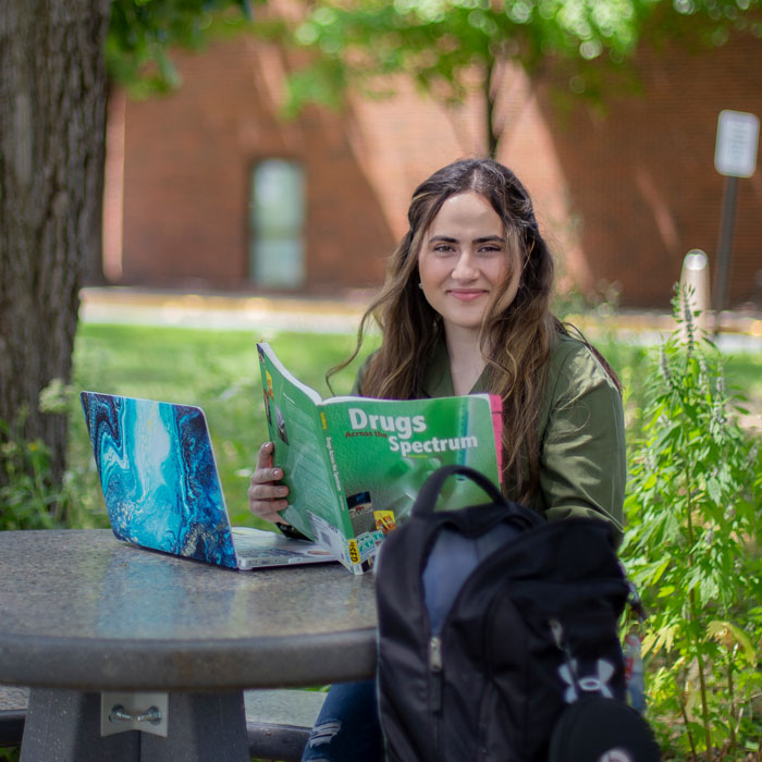College student seated at an outdoor campus table, reading a sociology textbook beside a laptop and backpack, with trees and a brick academic building behind.