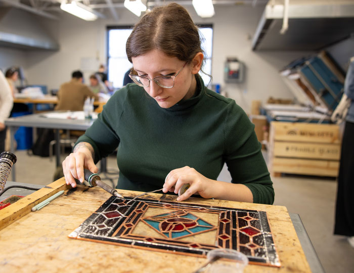 Student soldering pieces of a stained‑glass panel during a hands‑on studio art class at a workbench.
