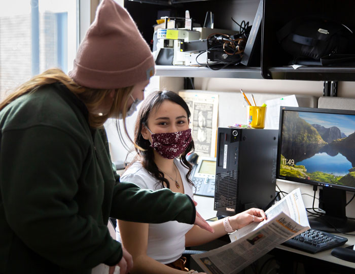 Two students working together at a desk in a campus office, reviewing a printed newspaper next to a desktop computer and monitor