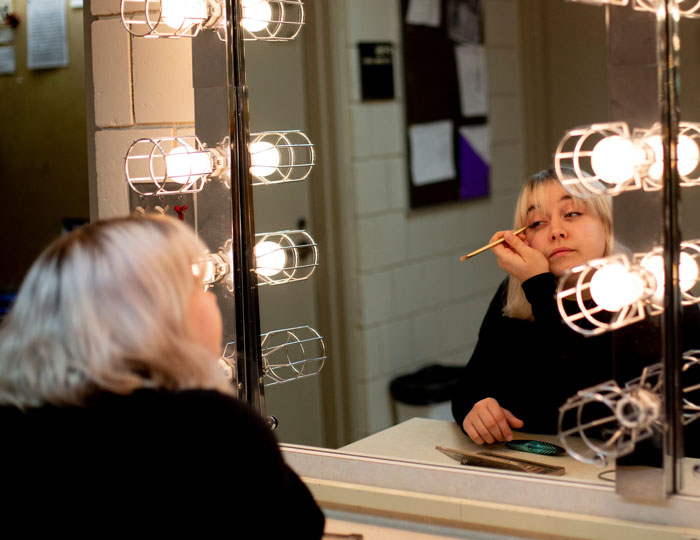 Student applying stage makeup at a lighted theater dressing room mirror.