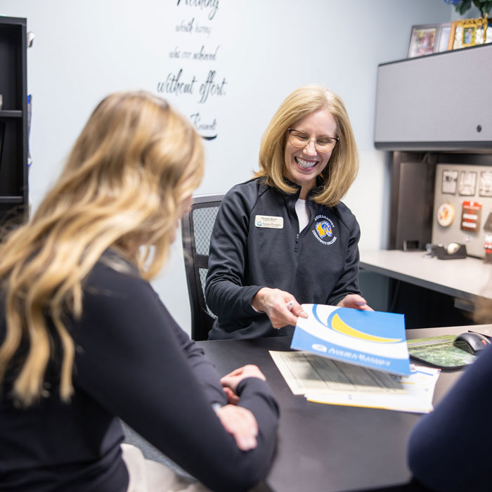 smiling staff member handing folder to student across a desk