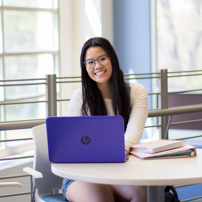 A smiling student typing on a purple laptop on campus with large windows in the background.