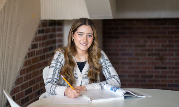 Student sitting at a round table on campus writing in a notebook with an open textbook.