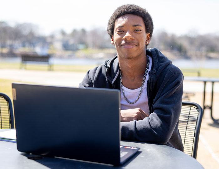 smiling student with arms crossed sitting at table outside with laptop