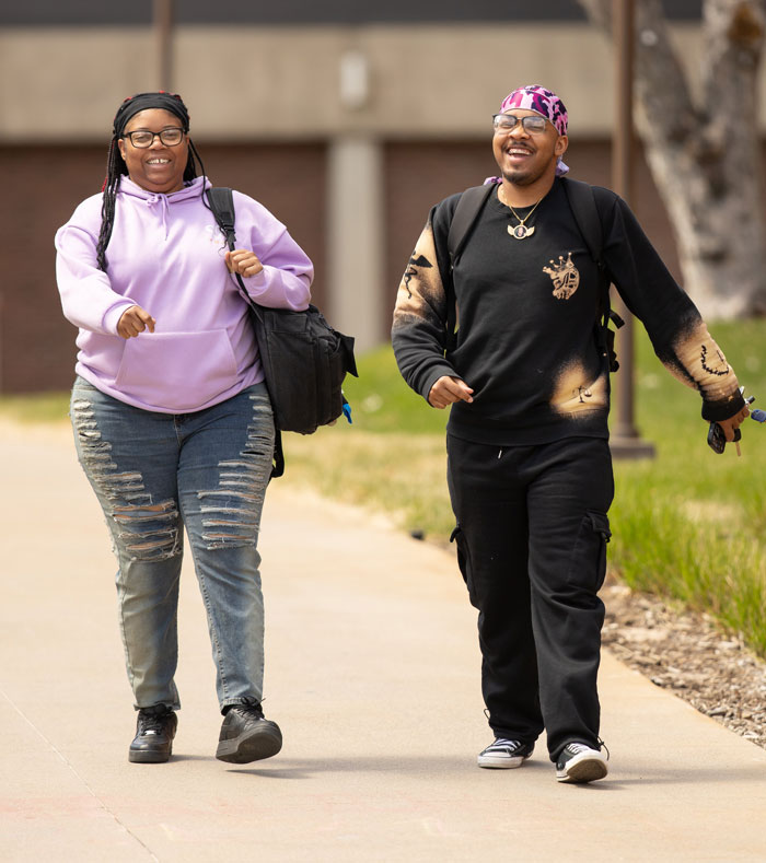 Two students walking side by side on a campus sidewalk, smiling and carrying backpacks.