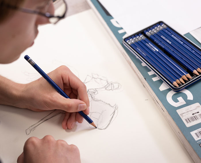 Art student uses graphite pencils to sketch a figure drawing on paper, illustrating foundational drawing skills taught in a college art class.
