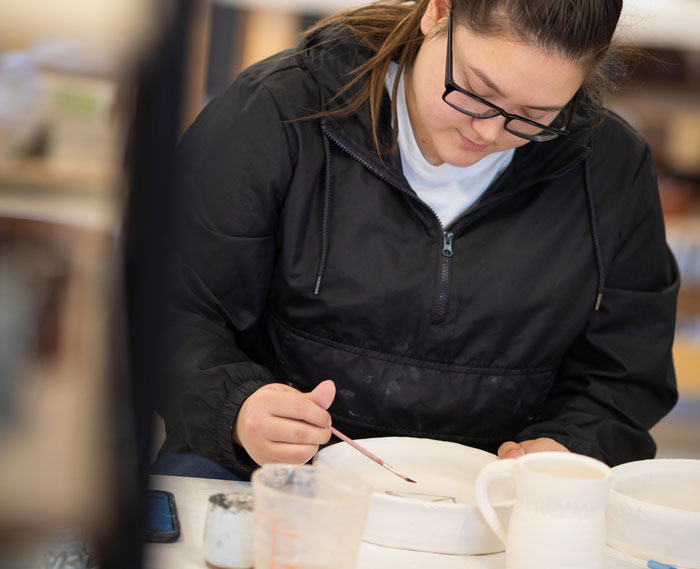 Art student applies glaze to a ceramic piece at a studio table, demonstrating hands-on ceramics coursework in a college visual arts program.