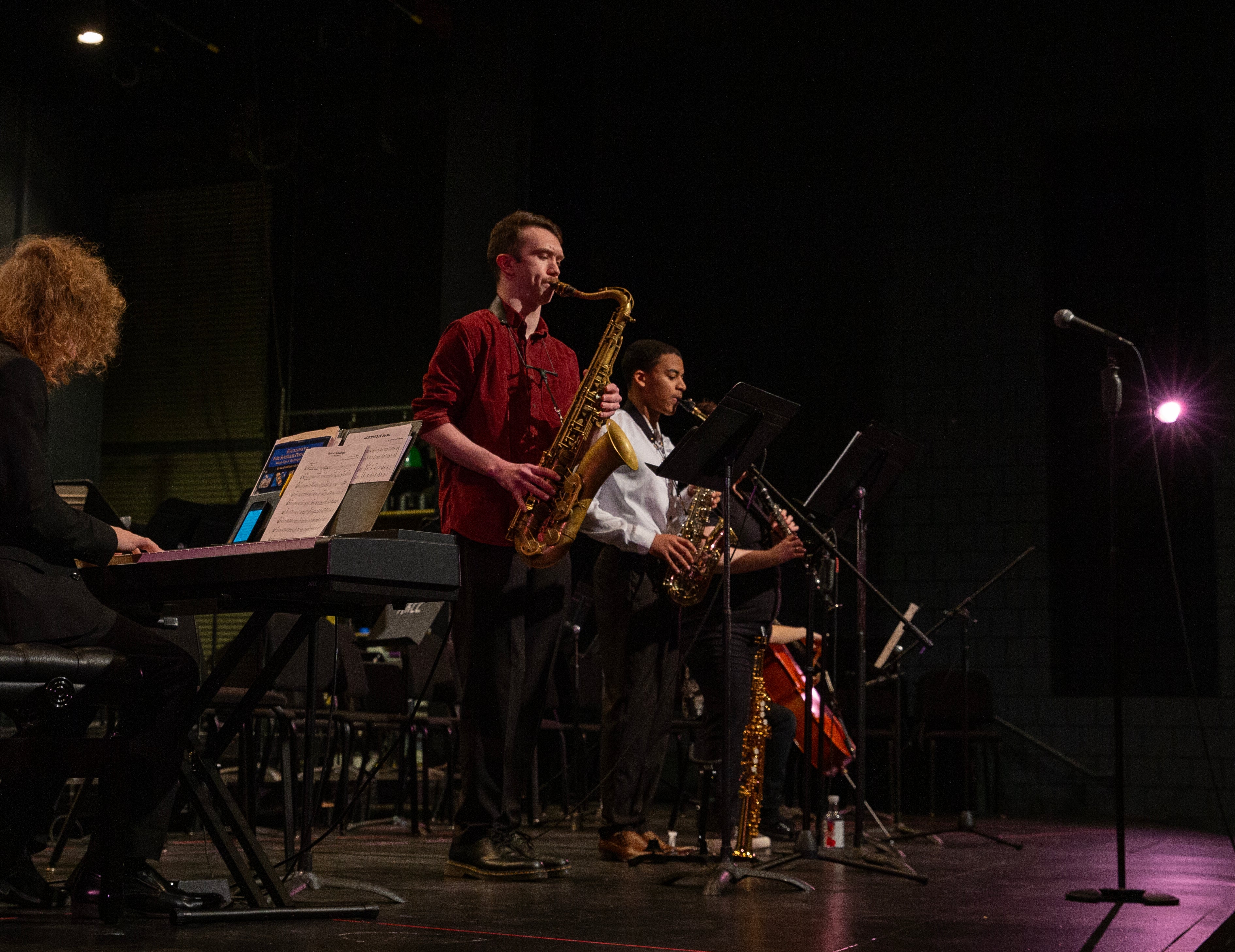 Two students playing the saxophone on a dimly lit stage with a purple spotlight to the right. One person is playing the piano. 