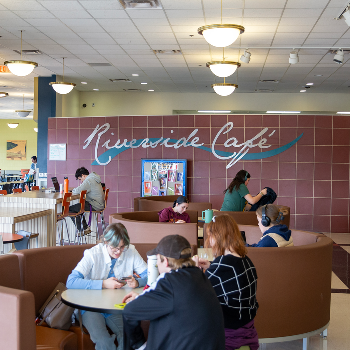 Riverside Cafe banner in the background with students sitting at tables talking, studying, eating, or looking at their phones.