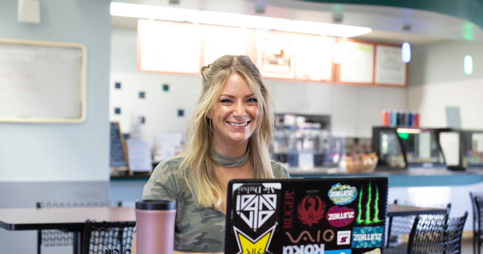 Cambridge Campus cafe with female student smiling at camera with laptop open in front of her.