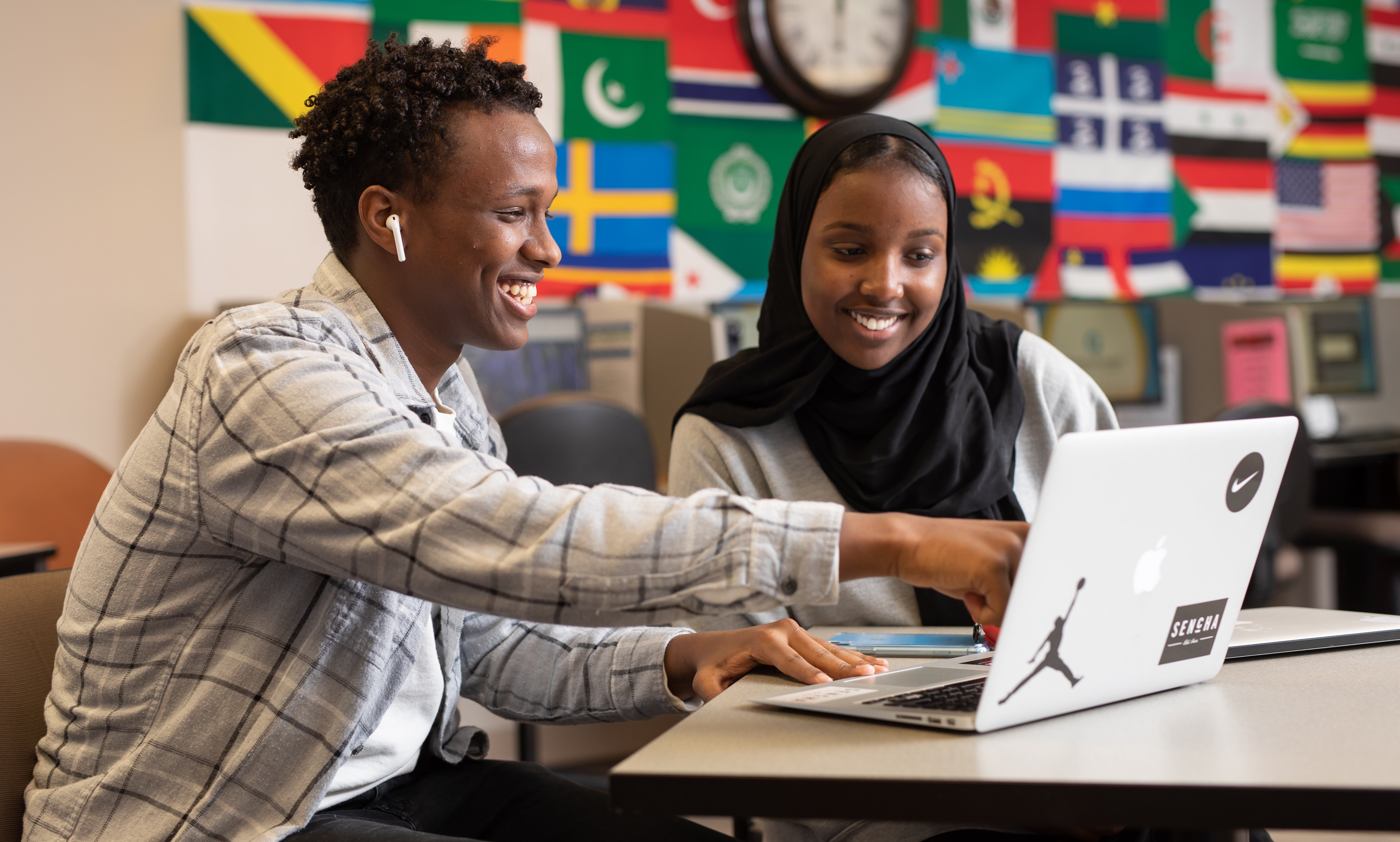 two students sitting at a table pointing at a computer smiling with a mural of flags in the background