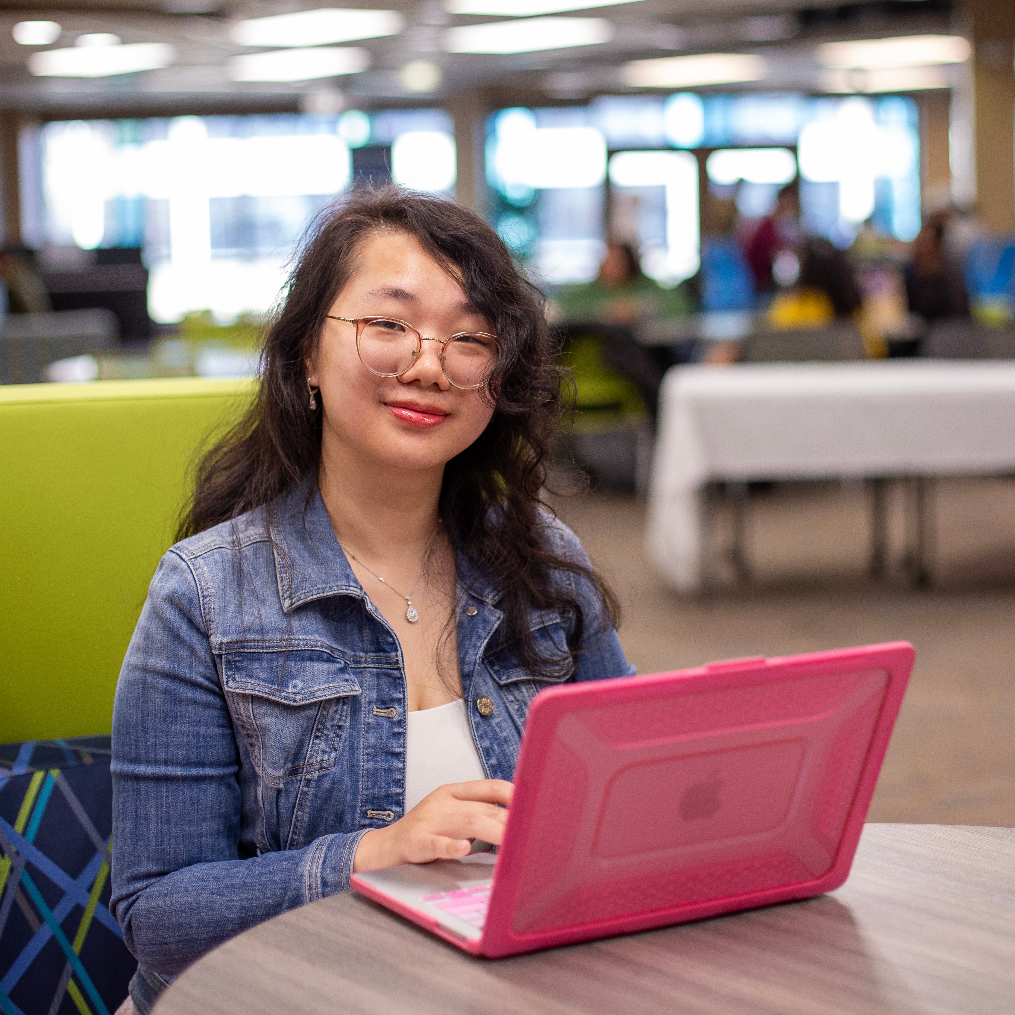 Student sitting at a table in the courtyard commons in the student center with her laptop in front of her, she is looking at the camera.