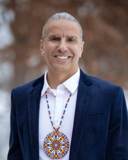 Image of man standing smiling and looking at the camera. The background is outside against a dark background. 