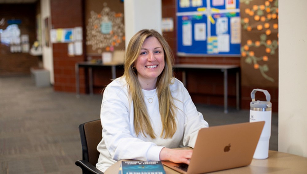 Individual seated in front of laptop, smiling at camera.