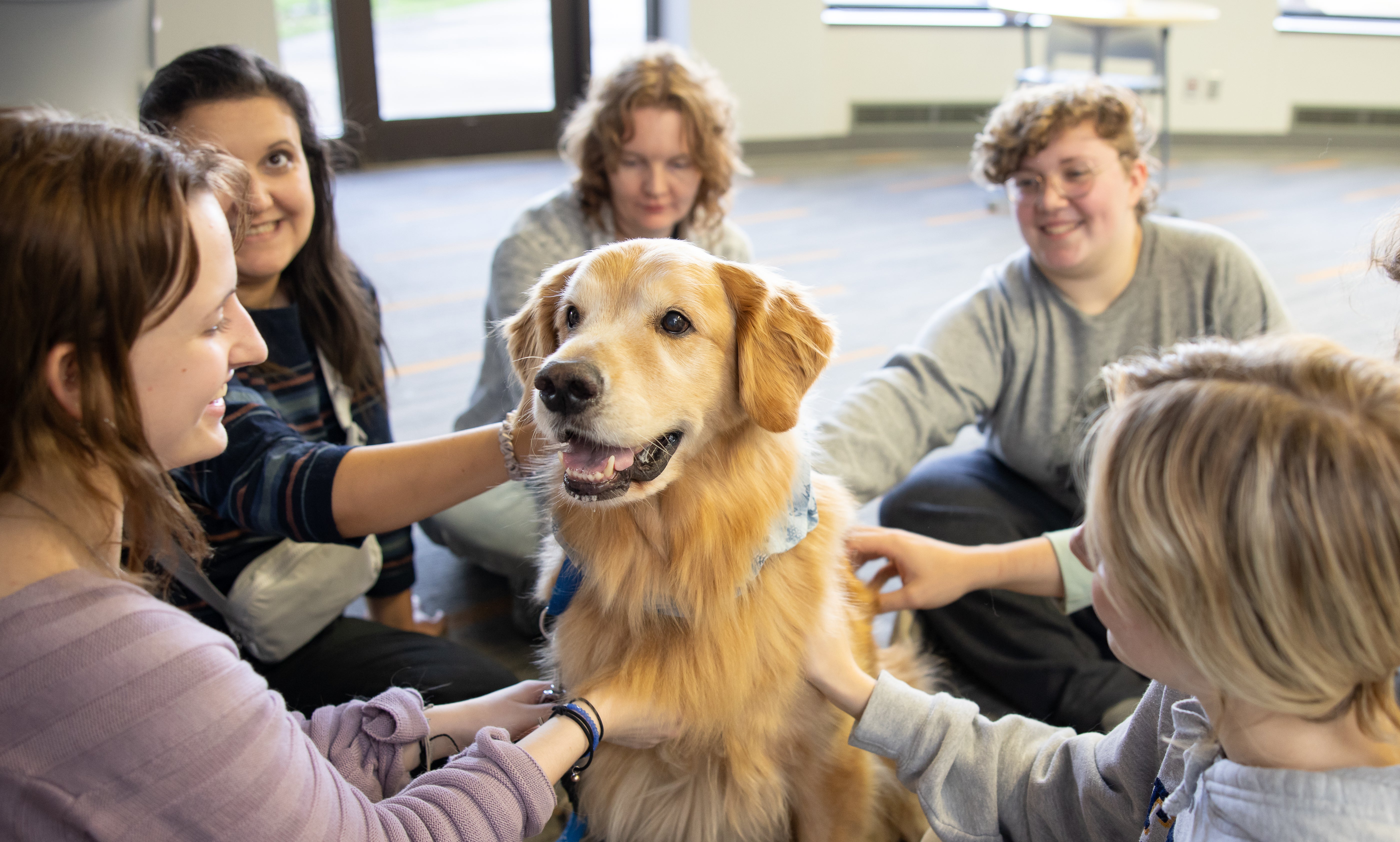 Five students sit in circle petting smiling at dog in center 
