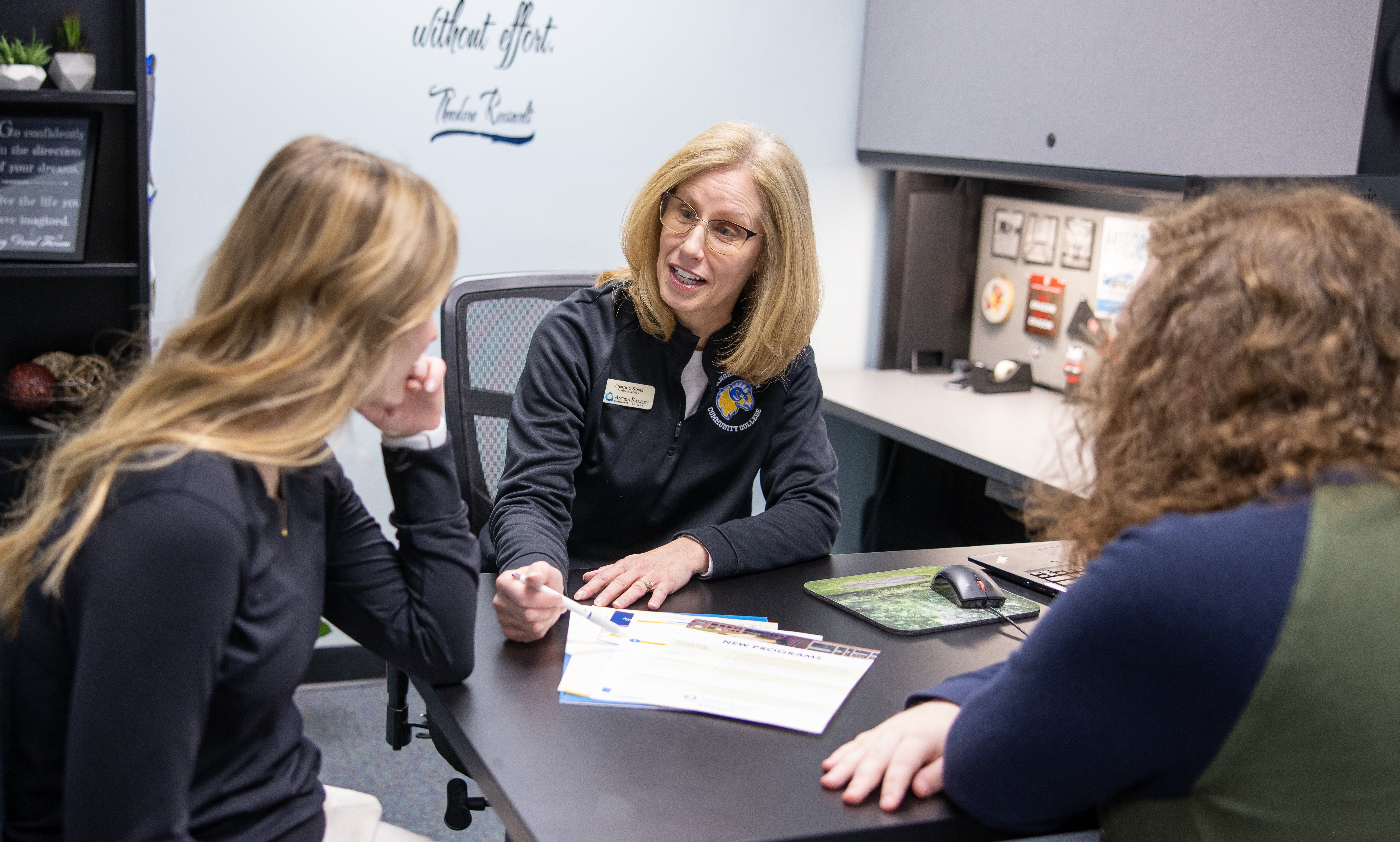 Female staff member sits at desk talking to two students sitting