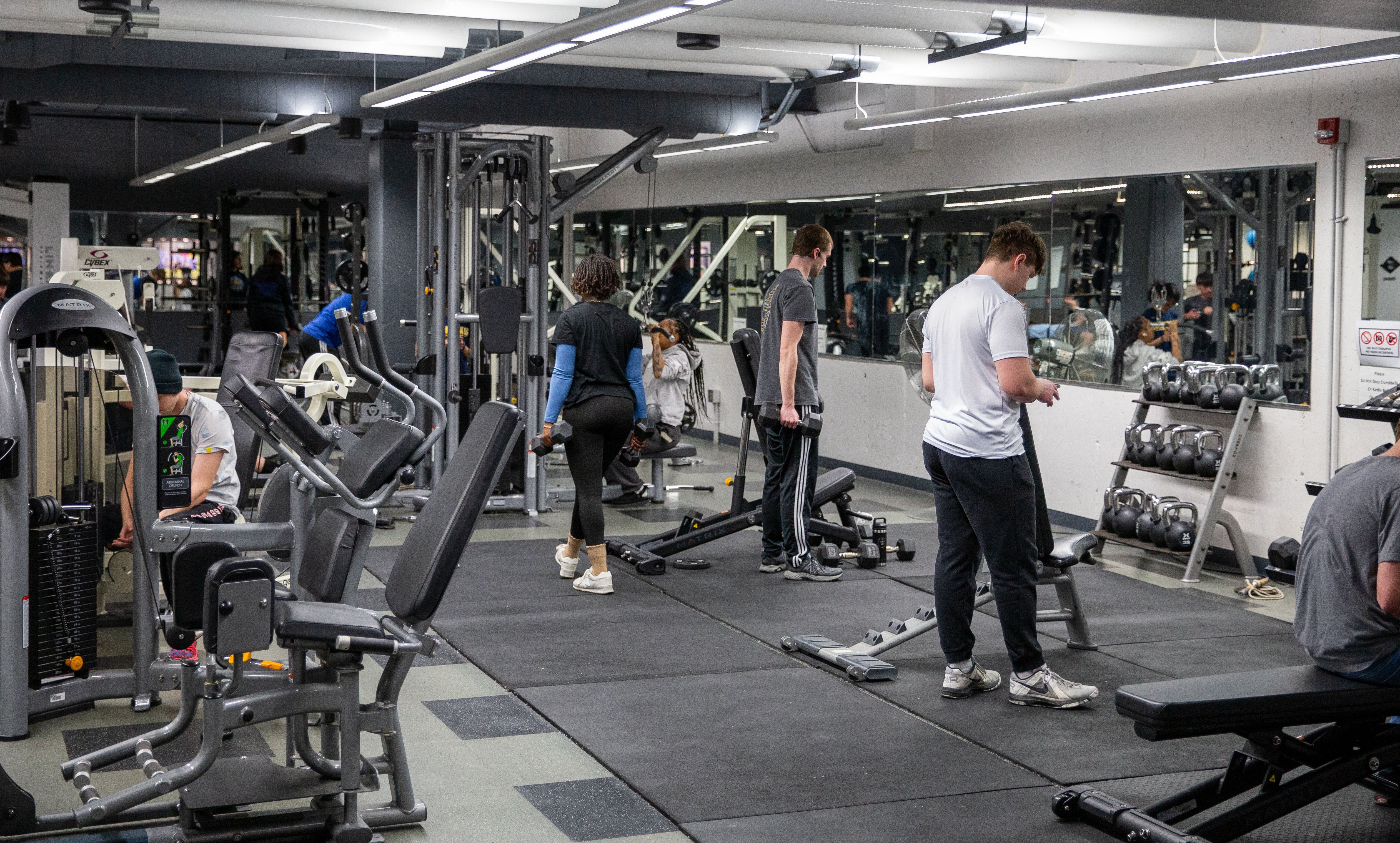 A gym with a variety of weights. There are three students holding weights with their backs to the camera.