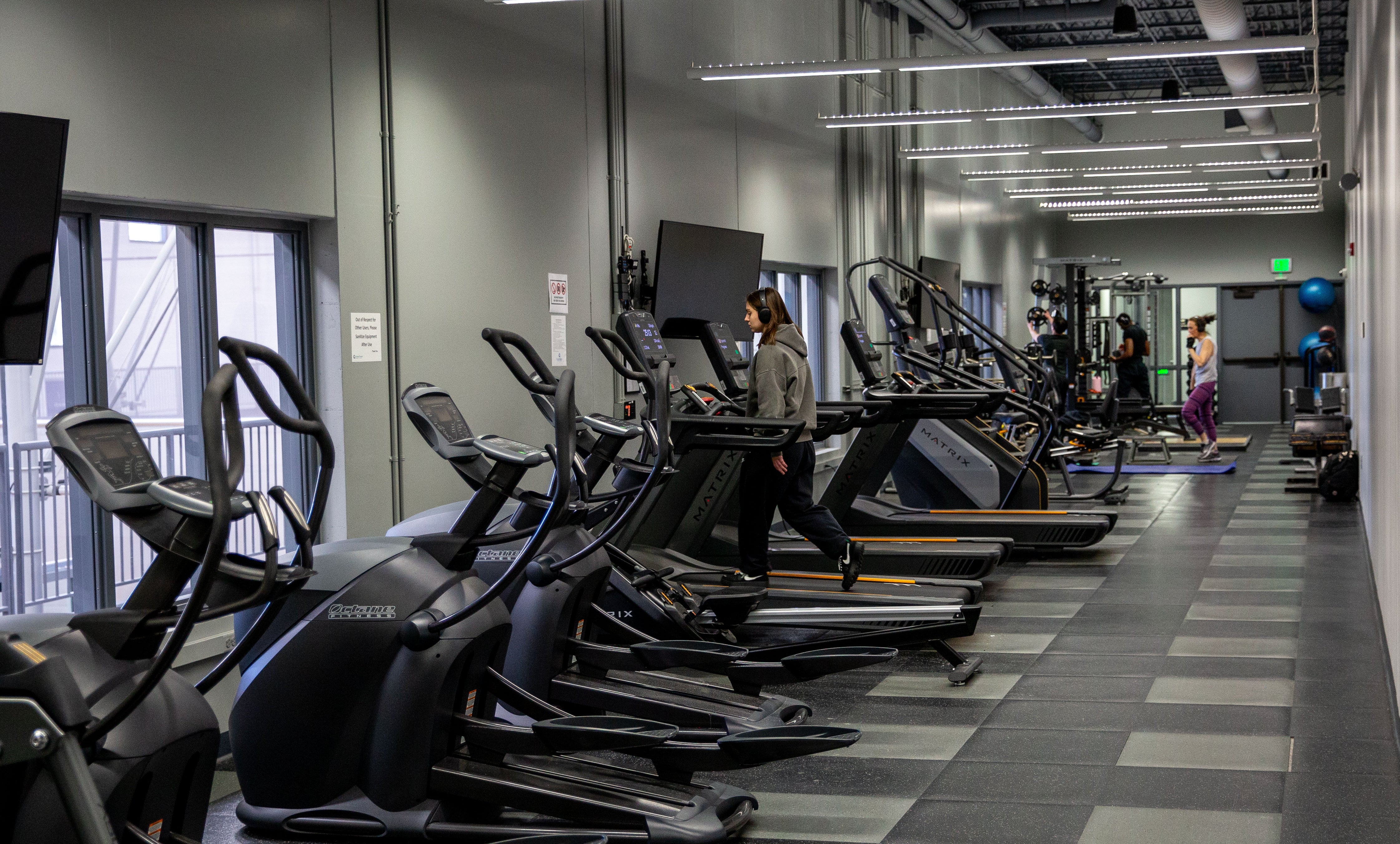 Mezzanie fitness center with a variety of tredmills, stair climber, and others. One person is walking on the tredmill and a person working out in the background.