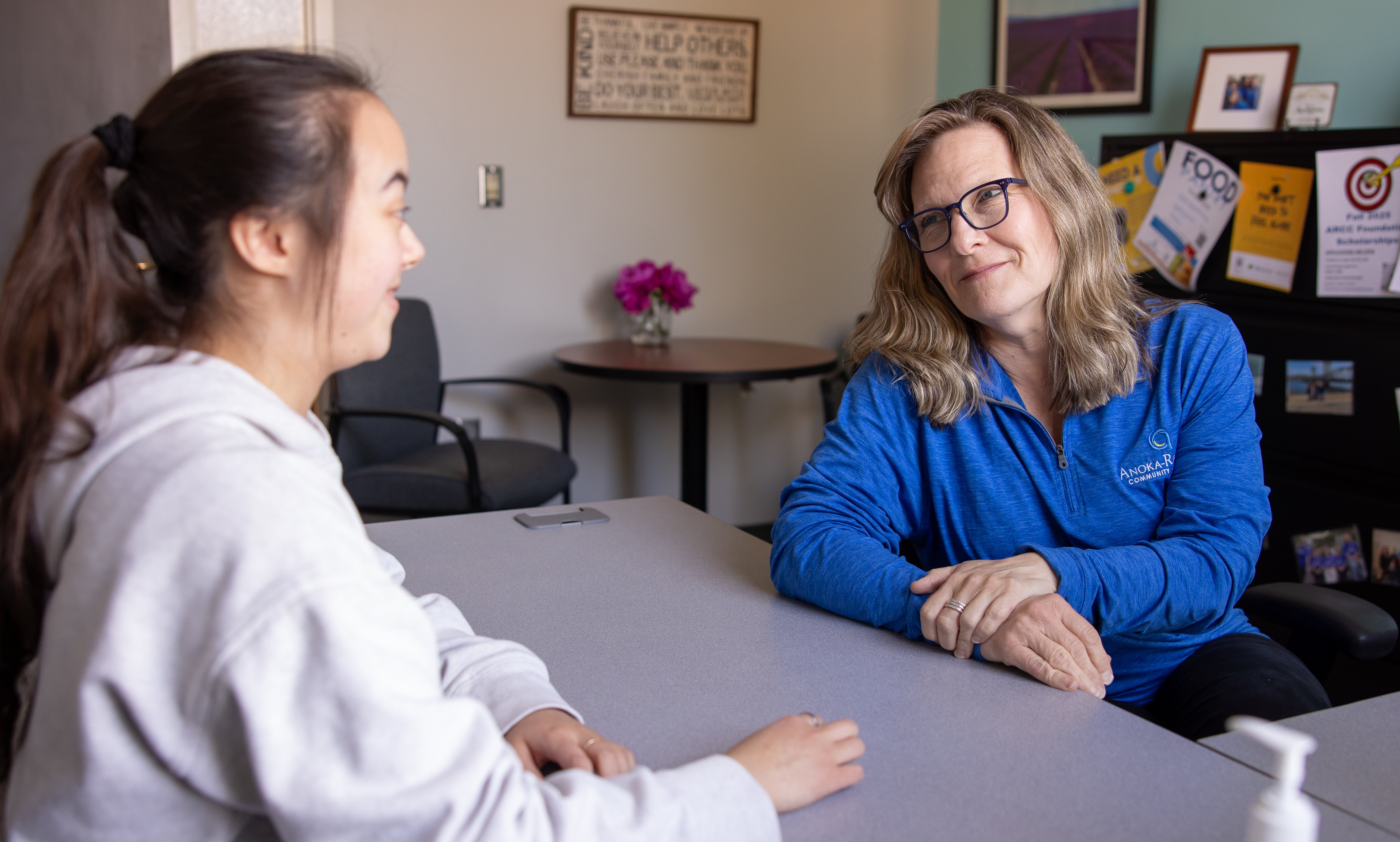 A female counselor wearing a blue shirt is sitting across the table from a female student wearing a white sweatshirt looking at each other